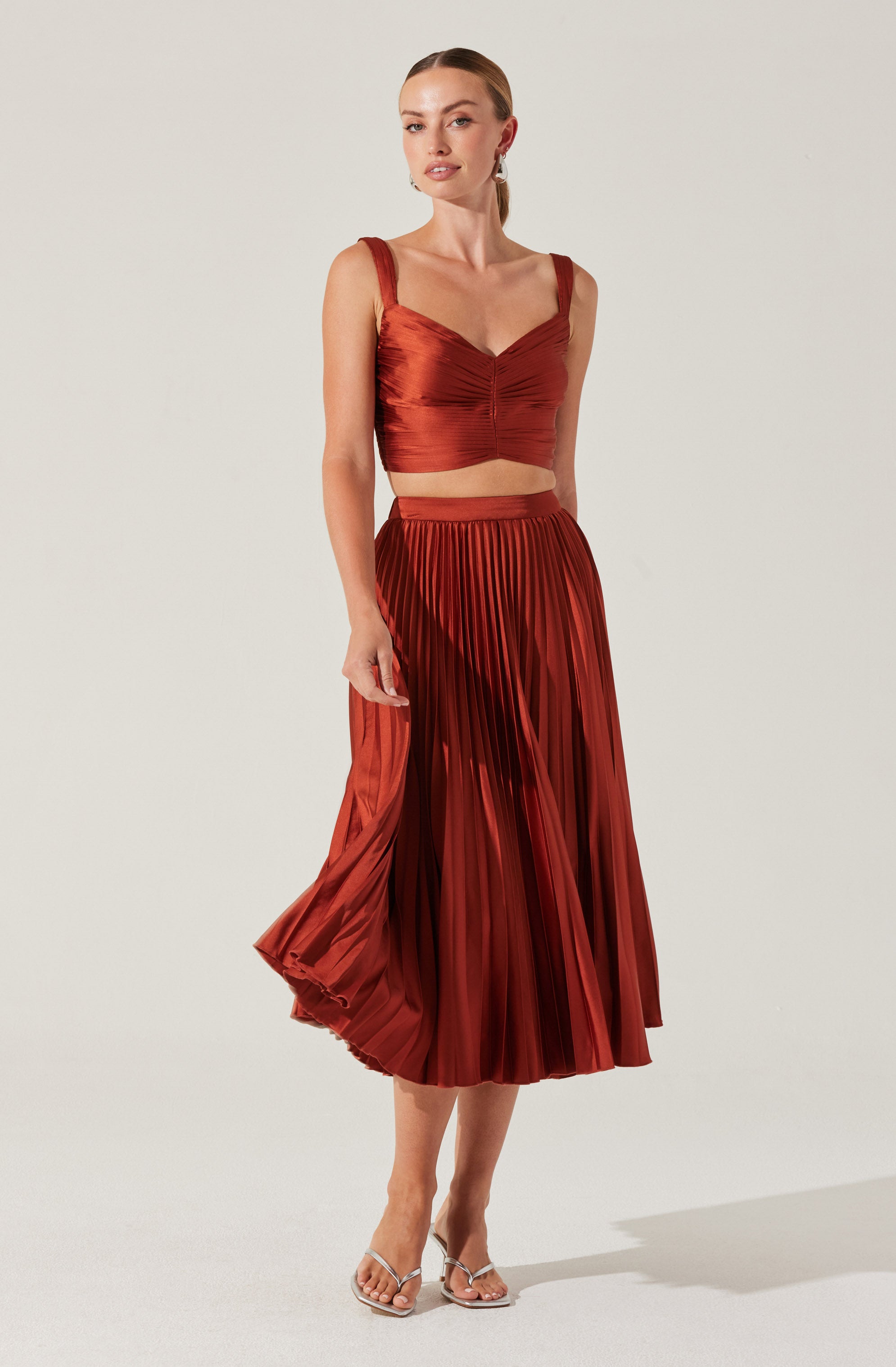 A woman is wearing a two-piece outfit in a shiny, rust-colored fabric. The top is a sleeveless, fitted crop top with wide straps and a sweetheart neckline, with ruching detail in the center. The skirt is a high-waisted, pleated midi-length skirt with a flowy, A-line shape. She is also wearing clear high-heeled sandals.