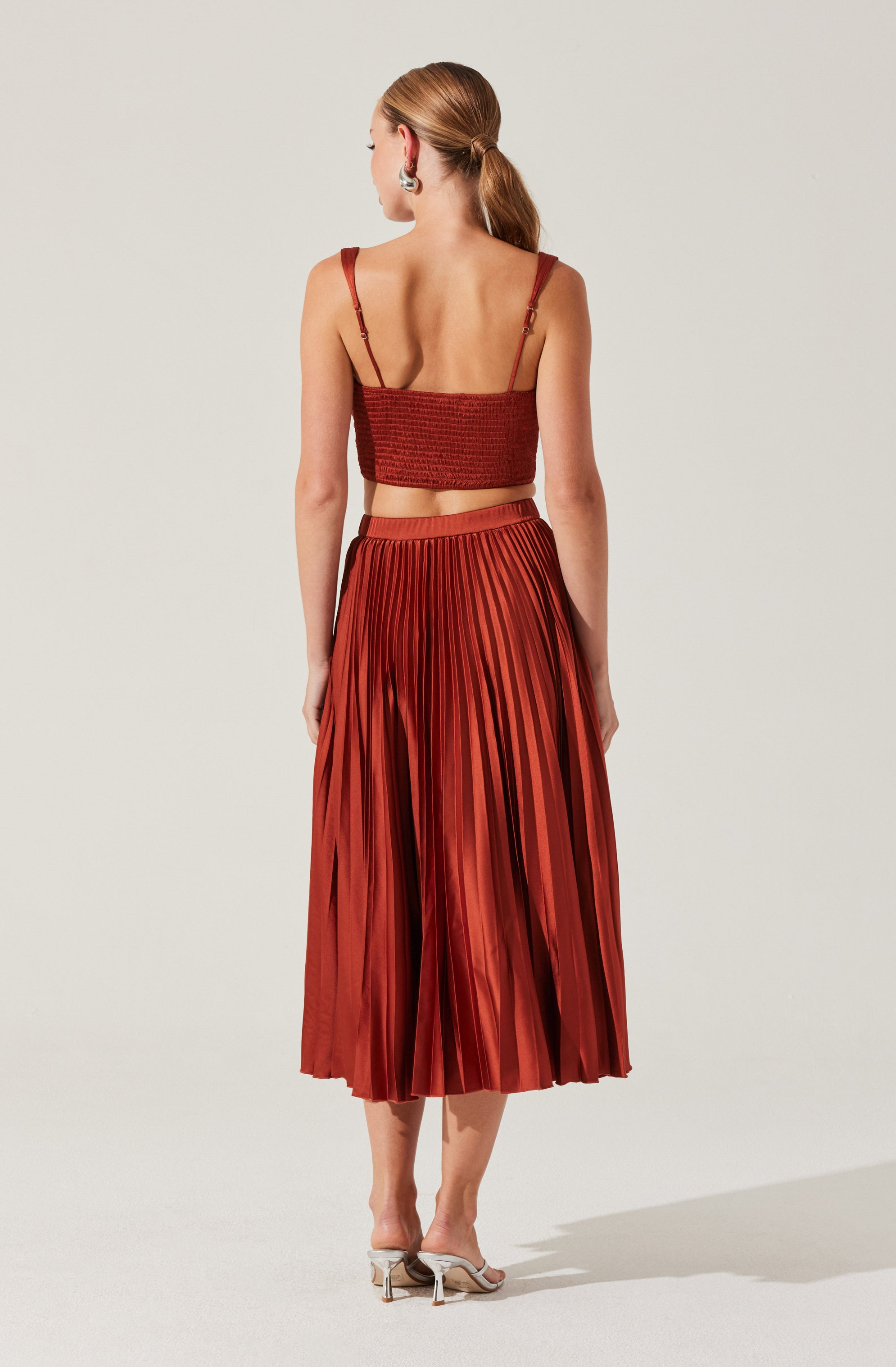 A woman is wearing a rust-colored, pleated midi skirt paired with a matching cropped top with spaghetti straps. She has her hair tied back in a low ponytail and is wearing statement earrings. She is also wearing open-toe, block-heeled shoes.