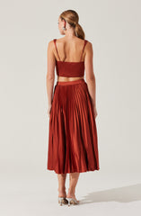 A woman is wearing a rust-colored, pleated midi skirt paired with a matching cropped top with spaghetti straps. She has her hair tied back in a low ponytail and is wearing statement earrings. She is also wearing open-toe, block-heeled shoes.