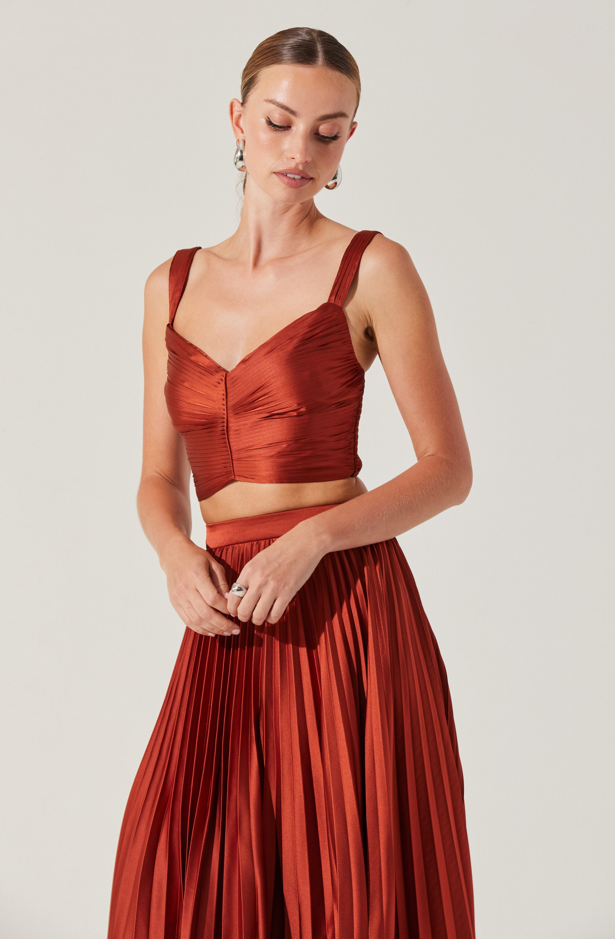 A woman is wearing a matching two-piece outfit in a shiny, rust-colored fabric. The top is a fitted cropped bustier with wide straps and a sweetheart neckline, featuring horizontal and gathering details. The skirt is a high-waisted, full-length, pleated design with a flowing silhouette.