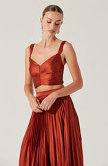 A woman is wearing a matching two-piece outfit in a shiny, rust-colored fabric. The top is a fitted cropped bustier with wide straps and a sweetheart neckline, featuring horizontal and gathering details. The skirt is a high-waisted, full-length, pleated design with a flowing silhouette.