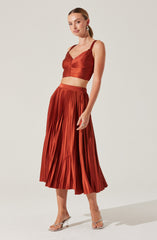 A woman is wearing a matching rust-colored outfit consisting of a crop top with a ruched bodice and adjustable shoulder straps, paired with a high-waisted, pleated midi skirt. She is also wearing clear, strappy high-heeled sandals and statement earrings.