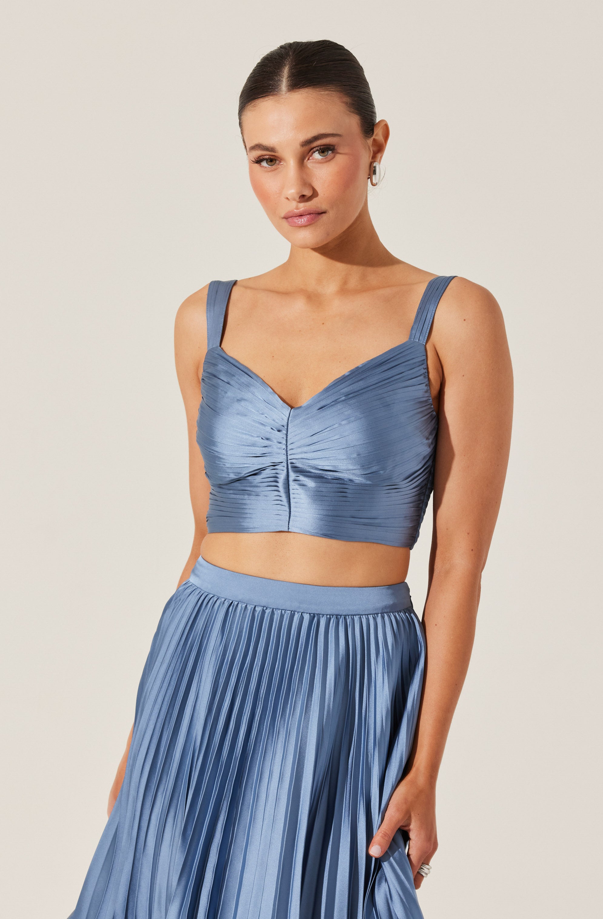 A woman wearing a two-piece outfit made of shiny, satin-like fabric in a soft blue color. The top is a fitted crop with wide straps, ruched detailing, and a V-shaped neckline. The matching skirt is high-waisted with a wide waistband and features vertical pleats throughout.