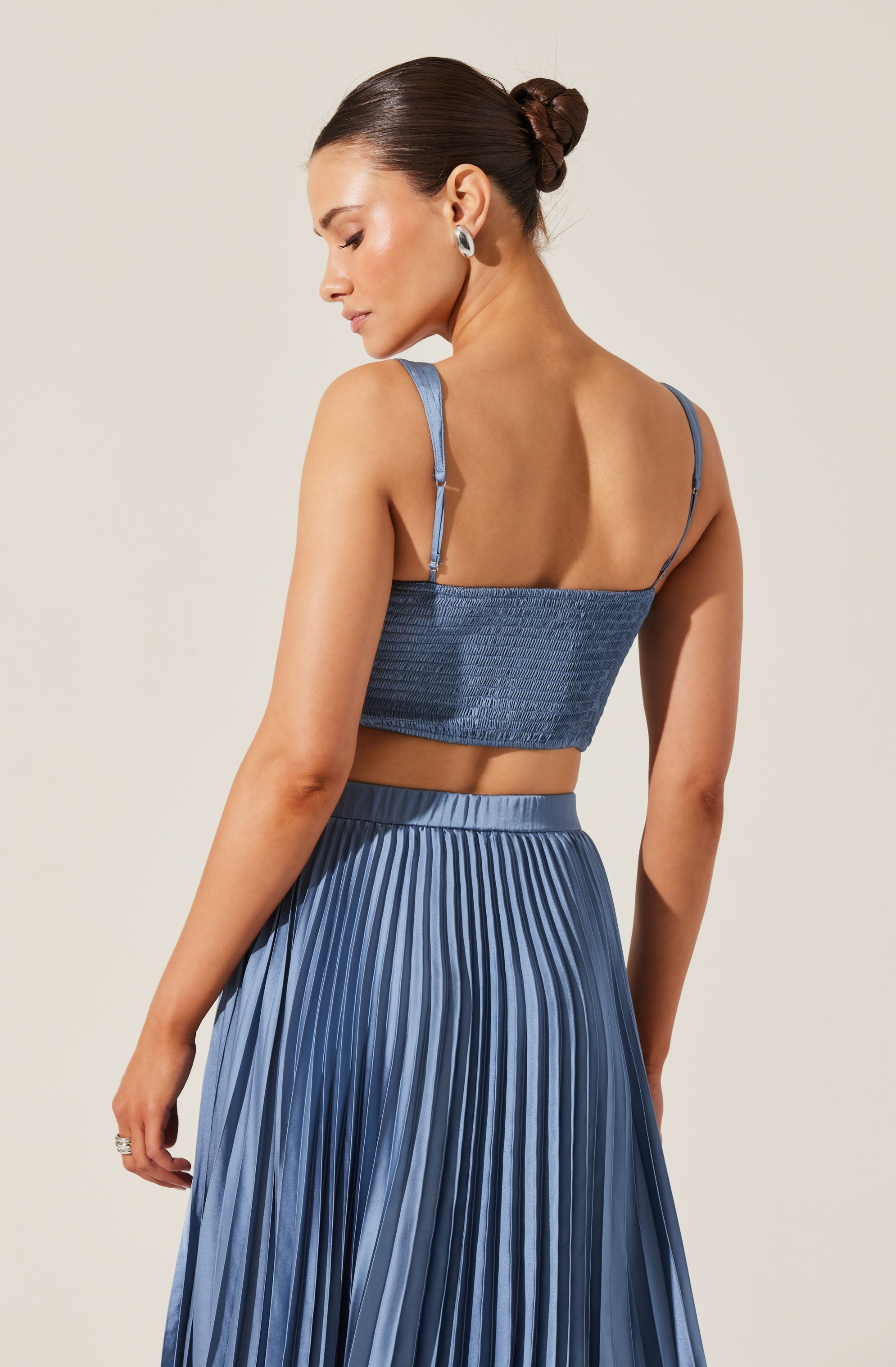 A woman is wearing a light blue, textured crop top with thin adjustable straps and a matching high-waisted pleated skirt in a darker shade of blue. The top has a smocked design, and the skirt features vertical pleats.