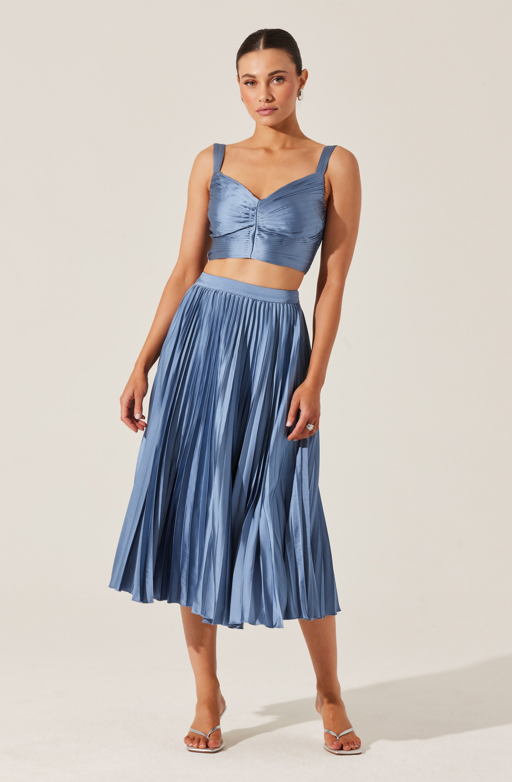 The outfit features a matching two-piece set in a shiny, silky blue fabric. The top is a cropped style with wide shoulder straps, a sweetheart neckline, and a ruched detail at the center. The skirt is a high-waisted, pleated midi-length design with a flowy silhouette. The model is paired the outfit with open-toed, clear high-heeled sandals.