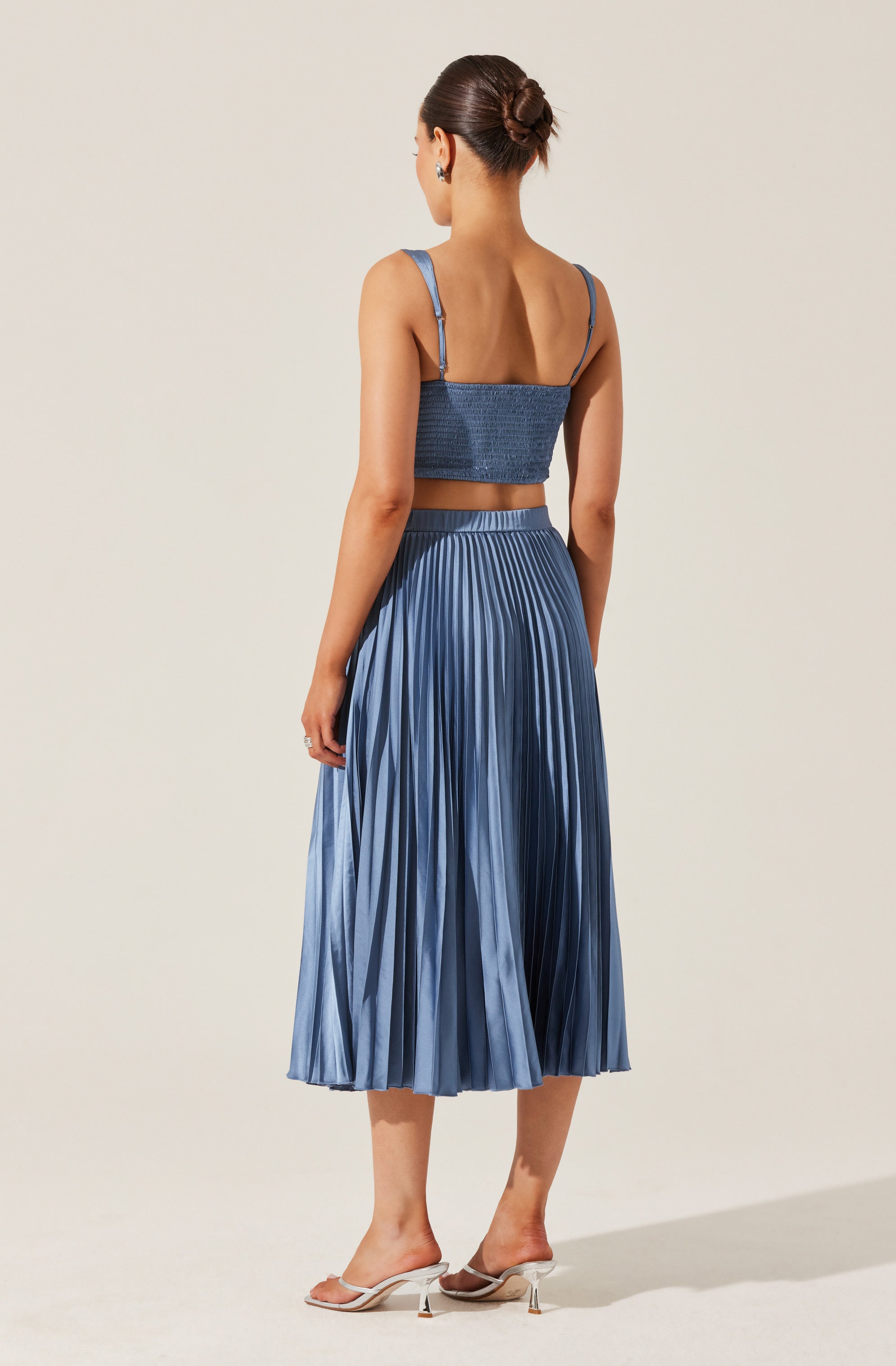 A woman is wearing a matching blue outfit consisting of a fitted, textured, sleeveless crop top with thin, adjustable shoulder straps and a high-waisted, metallic pleated midi skirt. She is also wearing open-toed, slip-on heels with transparent straps and a low heel. Her hair is styled in a neat, low bun.