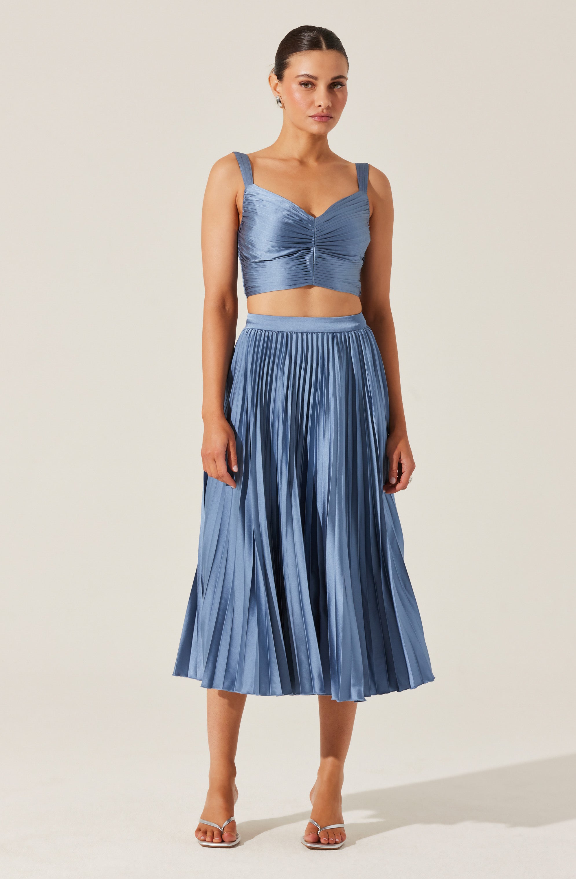 A woman is wearing a matching two-piece outfit in a shiny, satin-like fabric. The top has wide shoulder straps, a sweetheart neckline, and a ruched center detail. The high-waisted skirt is midi-length, pleated, and flows down to mid-calf. She is also wearing open-toe heels.