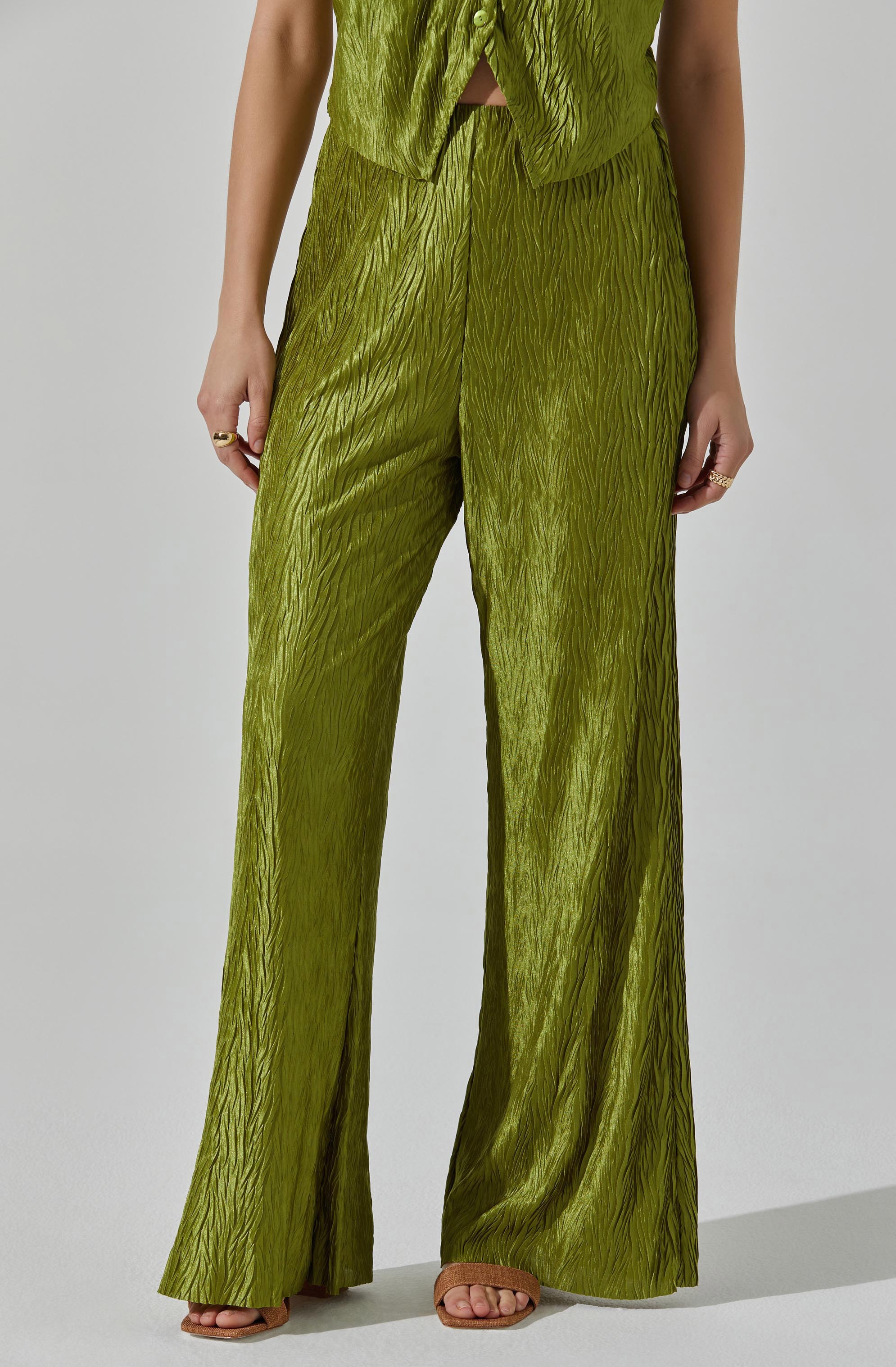 A person is wearing a matching set of shiny, textured olive green clothing. The top is sleeveless with buttons down the front and a small opening at the waist. The pants are high-waisted, wide-legged, and have a crinkled, shiny fabric that creates a textured appearance. The person is also wearing open-toed sandals and several rings on their fingers.