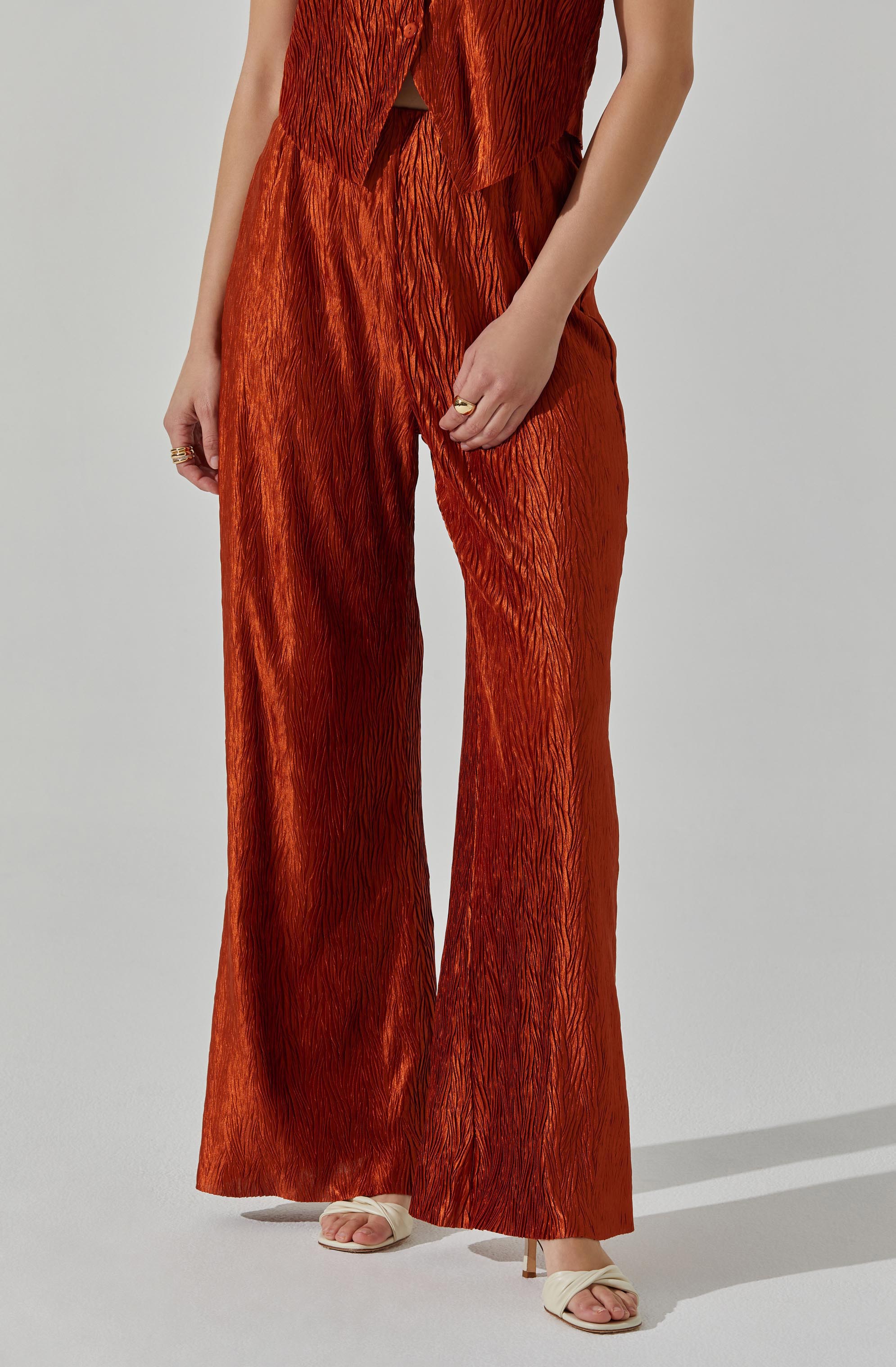The outfit consists of a sleeveless, rust-colored top with a textured, crinkled fabric and matching wide-leg pants made from the same textured material. The top features a front opening with a small slit, and the pants have a loose, flowing fit. The model is also wearing open-toed, pale-colored sandals with a block heel.