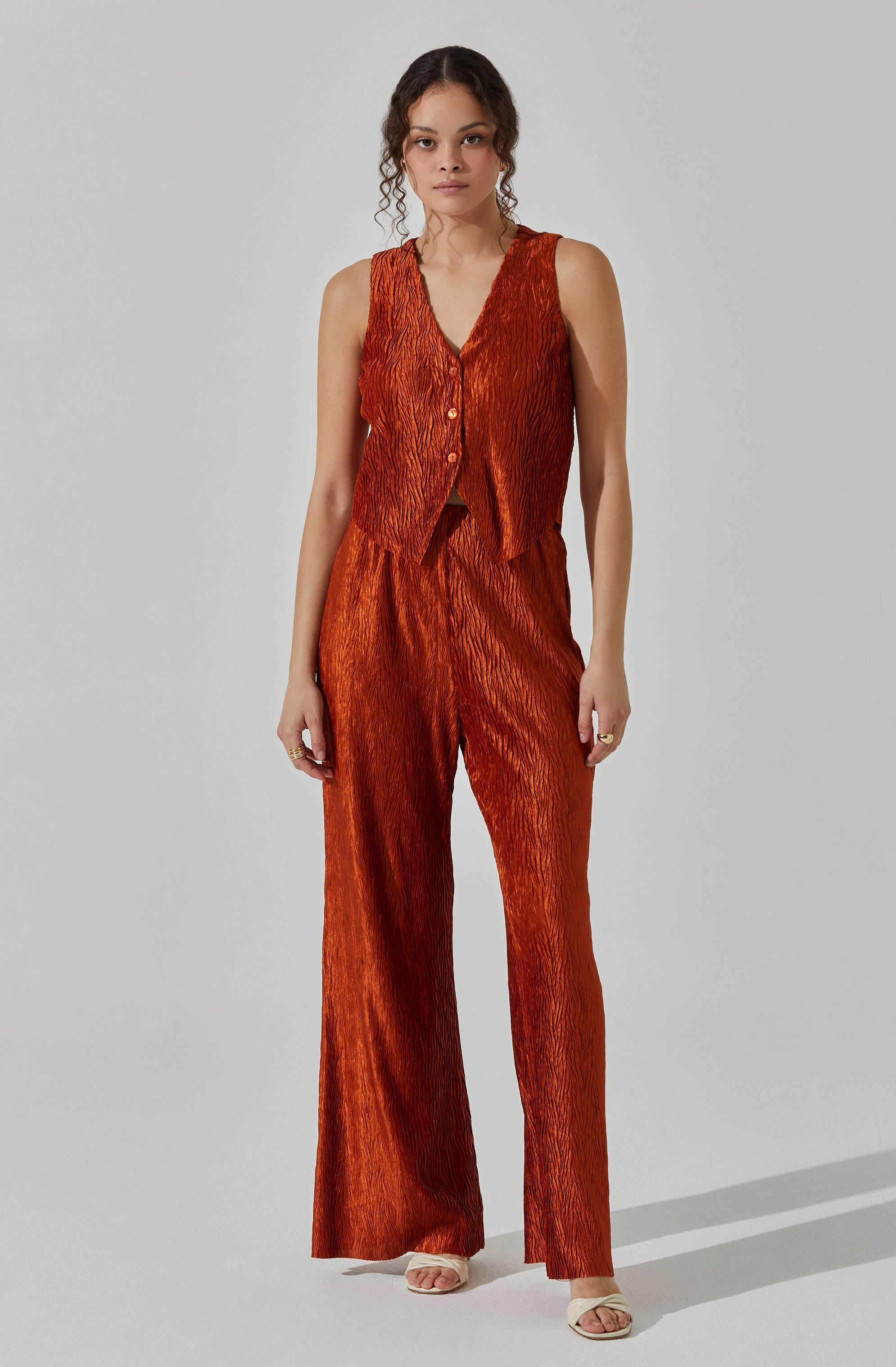 A woman is wearing a sleeveless, V-neck top with a textured, crinkled fabric in a rich, burnt orange color. The top features buttons down the front. She pairs it with matching high-waisted, wide-leg pants made of the same textured fabric.