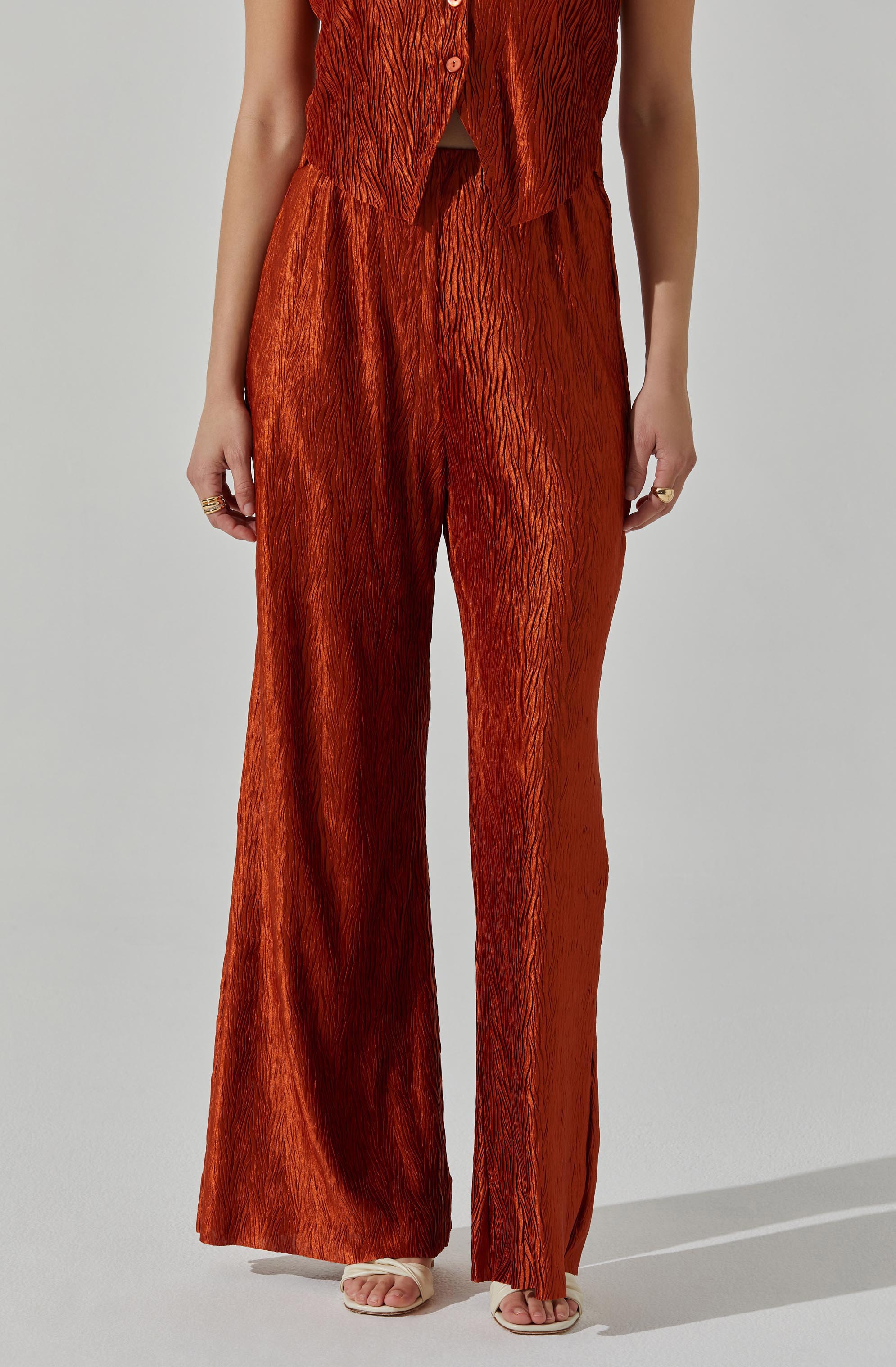 A person is wearing a matching orange-red outfit with a sleeveless top and wide-leg pants. The top appears to be a vest with buttons down the front, and the fabric has a textured, crinkled pattern. The pants are high-waisted and flow loosely to the floor, sharing the same textured fabric.