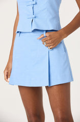 A person is wearing a light blue sleeveless blazer with decorative bows on the front and a matching light blue mini skirt with a front pleat. The individual is accessorizing with multiple rings on their fingers and a light blue ring on their wrist.