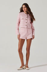 A woman is wearing a pink and white checkered cropped jacket with long sleeves and a round neckline, featuring decorative buttons down the front and on the pockets. She pairs it with matching pink and white checkered shorts. She is also wearing nude-colored heeled shoes with ankle straps.