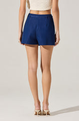 A person is wearing high-waisted, dark blue shorts with a tailored fit and side pockets. They are also wearing beige high-heeled shoes with ankle straps. The person has on a sleeveless, knitted, cream-colored crop top.
