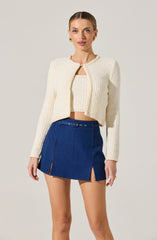 A woman is wearing a cream-colored, textured, long-sleeve, cropped cardigan with a round neckline and a matching tank top underneath. She pairs it with a high-waisted, dark blue denim mini skirt featuring small slits on both sides and a matching belt with a buckle.