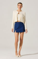 A woman is wearing a cream-colored, textured, long-sleeve, cropped cardigan with a round neckline and a small opening at the front. Underneath, she has a matching knit top. She pairs this with a high-waisted, dark blue mini skirt featuring two front slits and a small belt at the waist. She completes the outfit with light-colored pointed-toe heels.