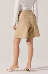 A person is wearing high-waisted beige shorts with a tailored fit, featuring back welt pockets and a centered front pleat. They are pairing the shorts with a white long-sleeve textured sweater. The outfit is completed with white open-toe heeled sandals with thin straps.