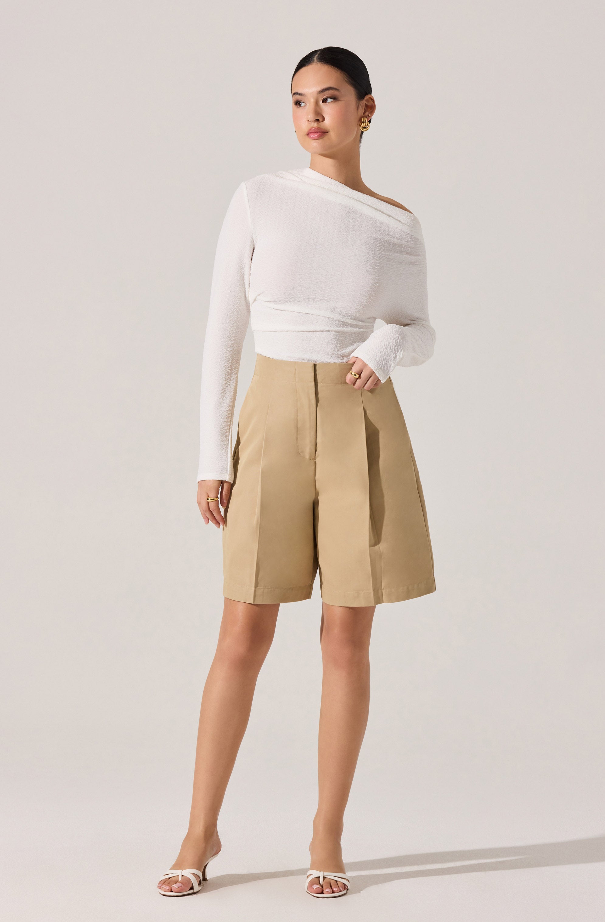 A woman is wearing a white, long-sleeved, asymmetrical sweater with a textured knit pattern, paired with high-waisted, beige knee-length shorts that have a wide-leg fit and pleats at the front. She is also wearing open-toed, strappy heels in a light color.