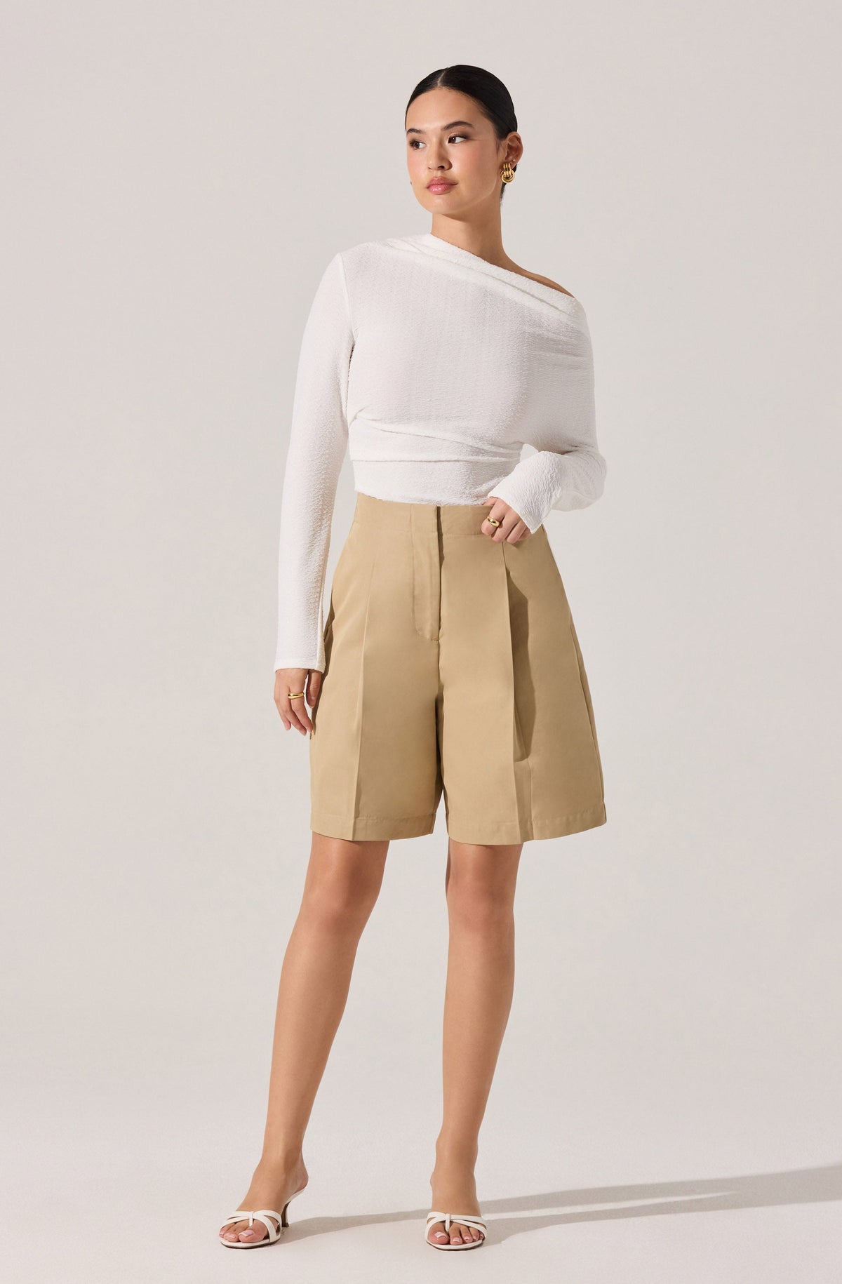 A woman is wearing a white, long-sleeved, asymmetrical sweater with a textured knit pattern, paired with high-waisted, beige knee-length shorts that have a wide-leg fit and pleats at the front. She is also wearing open-toed, strappy heels in a light color.