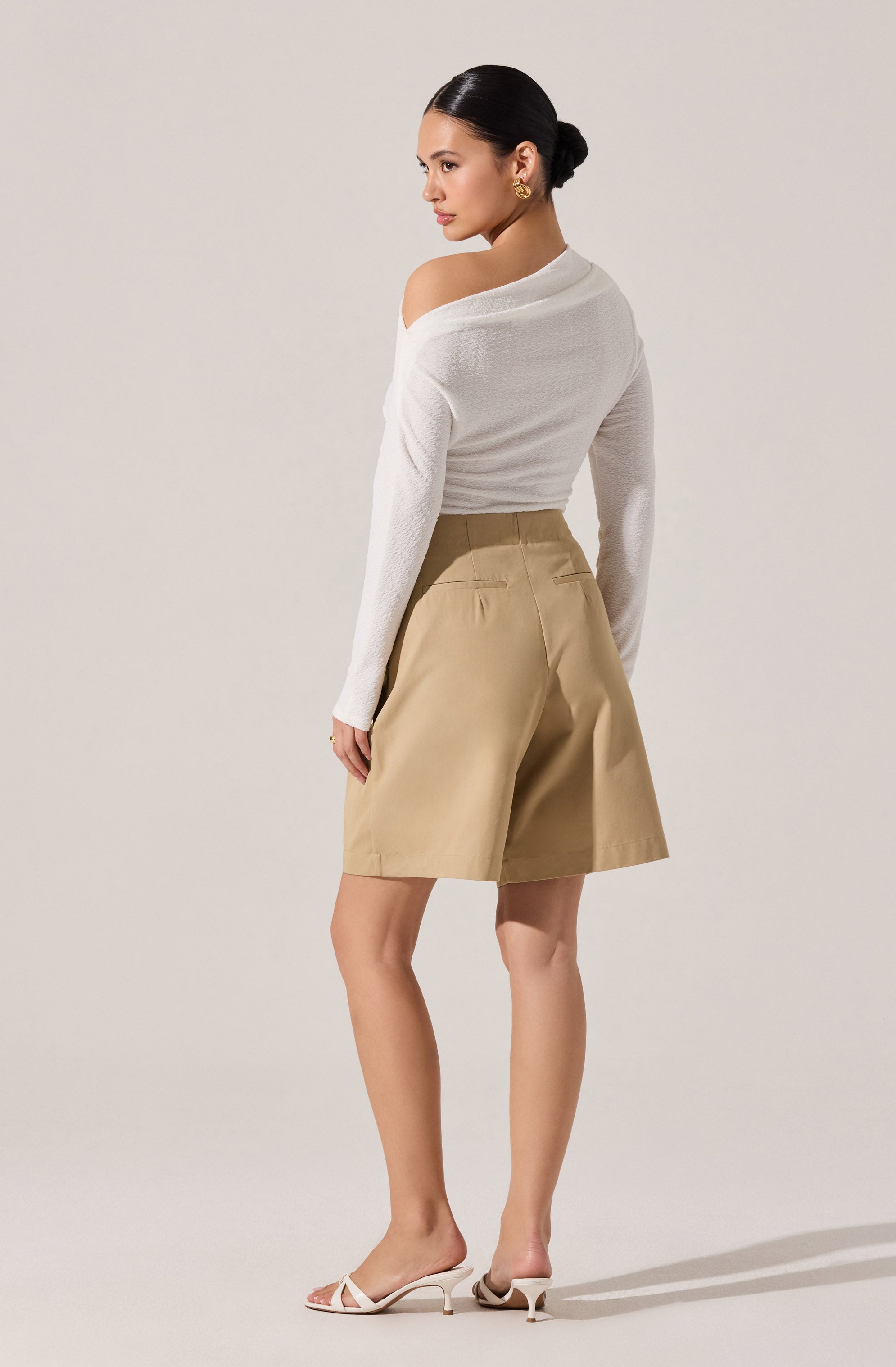 A woman is wearing a white, long-sleeved, off-the-shoulder sweater and beige high-waisted shorts with back pockets, paired with white high-heeled sandals.