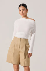 A woman is wearing a white, long-sleeved, off-the-shoulder sweater with a textured fabric, paired with beige high-waisted, wide-leg shorts that have pleats and a front zipper. She accessorizes with gold hoop earrings and a gold ring.