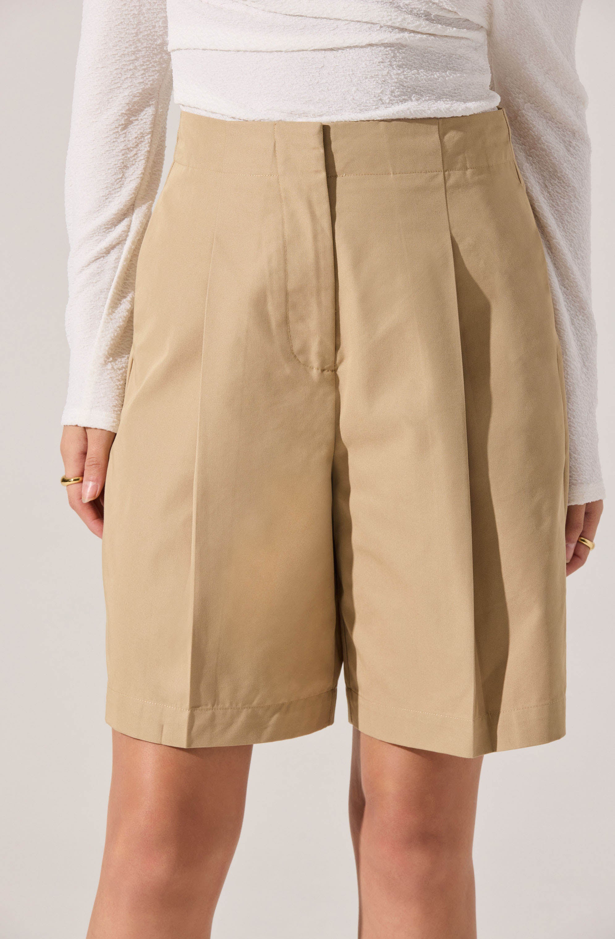 A person is wearing high-waisted beige shorts with a tailored fit, pleats at the front, and a wide waistband. They are also wearing a long-sleeved, white textured top tucked into the shorts.