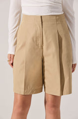 A person is wearing high-waisted beige shorts with a tailored fit, pleats at the front, and a wide waistband. They are also wearing a long-sleeved, white textured top tucked into the shorts.