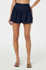 A woman is wearing high-waisted navy blue shorts with a gathered, ruffled design and a wide waistband. She is also wearing black strappy high-heeled sandals.
