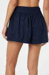 The image shows a person wearing navy blue shorts with a wide waistband and gathered fabric at the back. The shorts have a loose fit and are made of a lightweight fabric. The person is standing with their back to the camera, and the background is plain and light-colored.