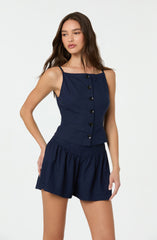 A young woman is wearing a navy blue sleeveless top with thin shoulder straps and a row of black buttons down the front. She pairs it with matching navy blue shorts that have a gathered, ruffled hem.