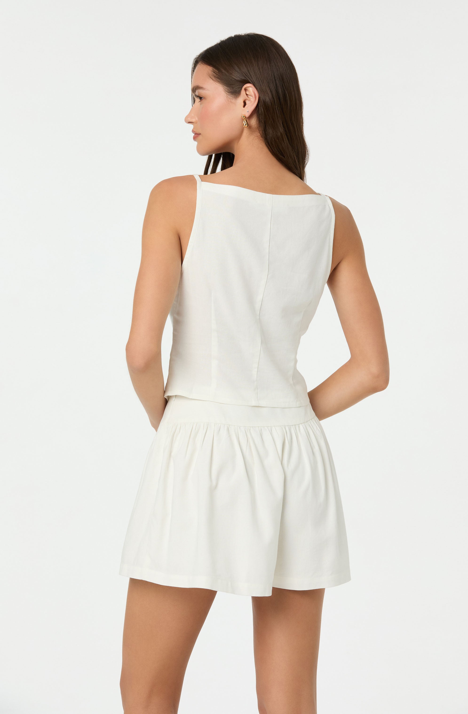 A woman is wearing a sleeveless, off-white top with wide shoulder straps and a straight neckline. The top appears to be made of a lightweight fabric and fits snugly around her torso. She is also wearing matching off-white high-waisted shorts with an elastic waistband, gathered fabric, and a relaxed fit.
