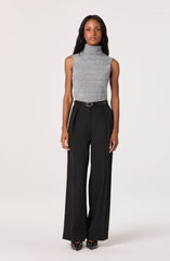A woman is wearing a sleeveless, high-neck, grey knit top paired with high-waisted, wide-leg black trousers. The trousers have a belt and feature a pleated front design. She completes her outfit with patterned high-heeled shoes.