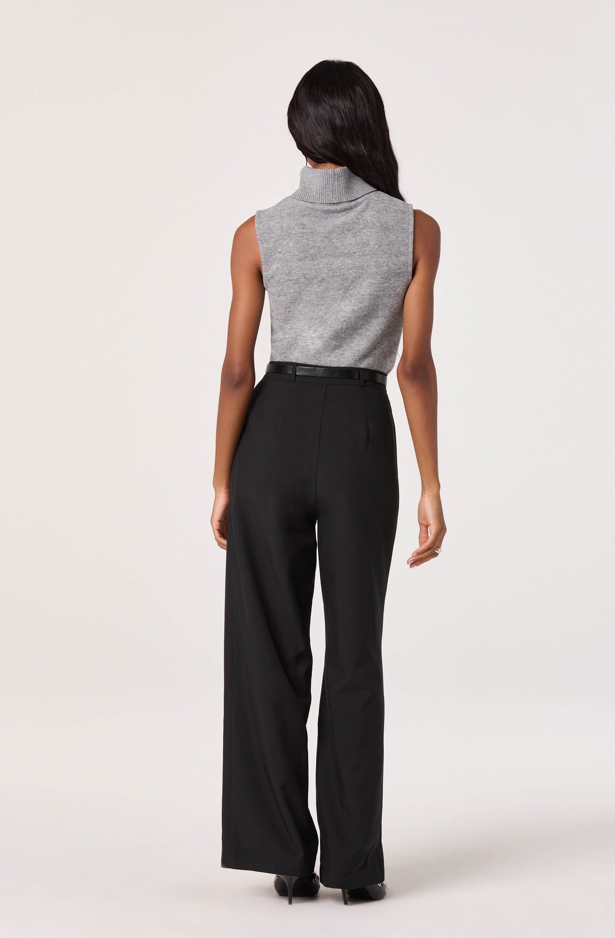 A woman is wearing a sleeveless, gray turtleneck sweater with a high collar that fits snugly around her neck. She is also dressed in high-waisted, black wide-leg trousers that reach the floor, paired with black shoes.