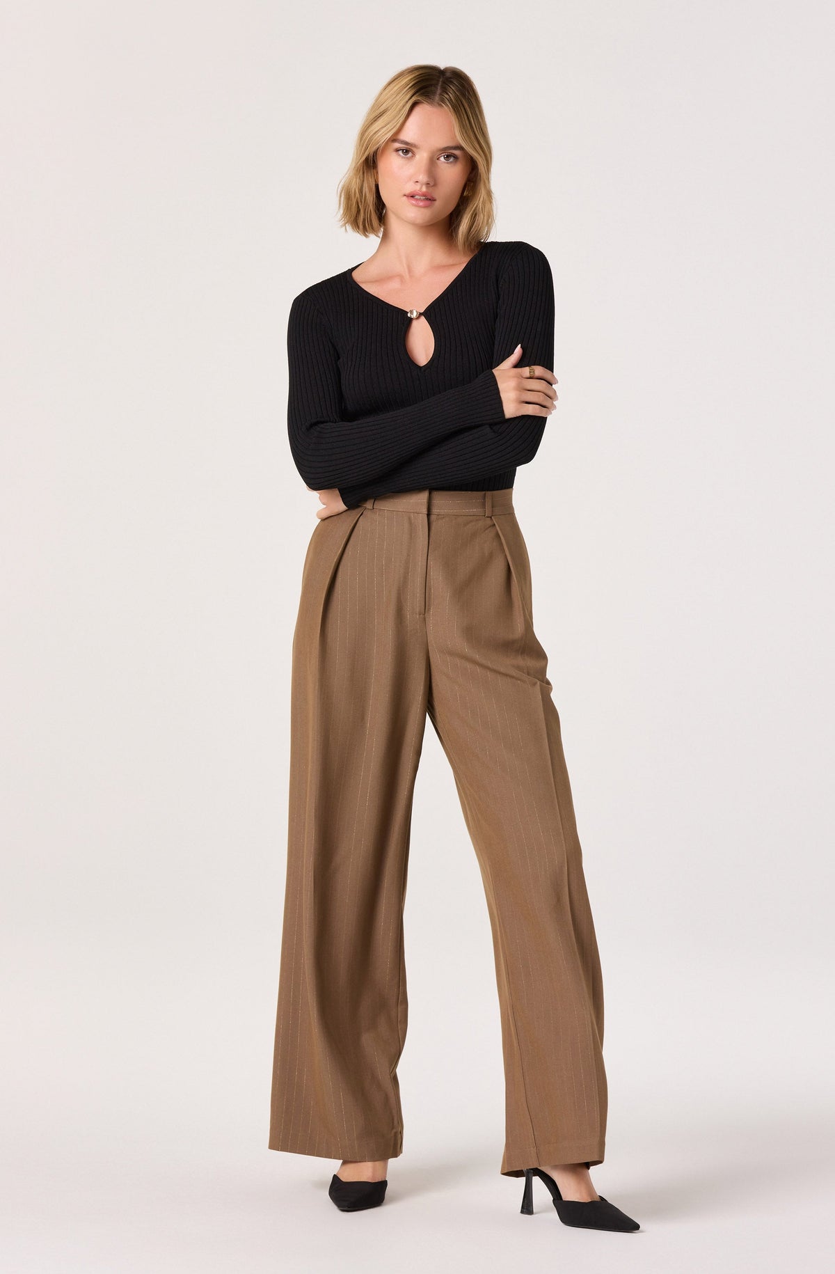 A woman is wearing a fitted, long-sleeved, black ribbed sweater with a small keyhole opening secured by a decorative clasp at the neckline. She is also wearing high-waisted, wide-leg tan trousers with a subtle pinstripe pattern.