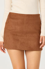 A woman is wearing a high-waisted, A-line brown suede mini skirt. She is also wearing a light beige, long-sleeve sweater tucked into the skirt. Her right hand is resting gently at her side, and her nails are painted with a light polish.