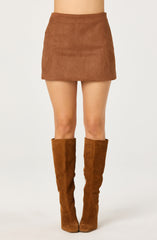 A person is wearing a brown suede mini skirt that is fitted at the waist and ends mid-thigh. They are also wearing tall, brown suede knee-high boots with a slight heel. The background is plain and light-colored.