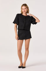 A young woman with shoulder-length blonde hair is wearing a black textured outfit. The top is a short-sleeved, boxy jacket with puffed sleeves, featuring small bows down the front. The matching skirt is short, fitted, and has the same textured fabric. She is also wearing black shoes with bows on the front and gold rectangular earrings.