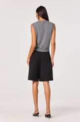 A woman is wearing a sleeveless, gray, knitted top with padded shoulders. She pairs it with high-waisted, black knee-length shorts. Her footwear consists of black strappy heels with thin stiletto heels.