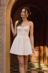A woman is wearing a short, cream-colored dress with thin spaghetti straps and a fitted, textured bodice. The dress has a flared, pleated skirt that falls above her knees. She also has large, round earrings and a white hair accessory.