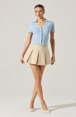 A woman is wearing a light blue short-sleeved button-up sweater with a textured, open-knit pattern and a collar. She pairs it with a beige, high-waisted, pleated mini skirt. She completes the look with beige pointed-toe heels.