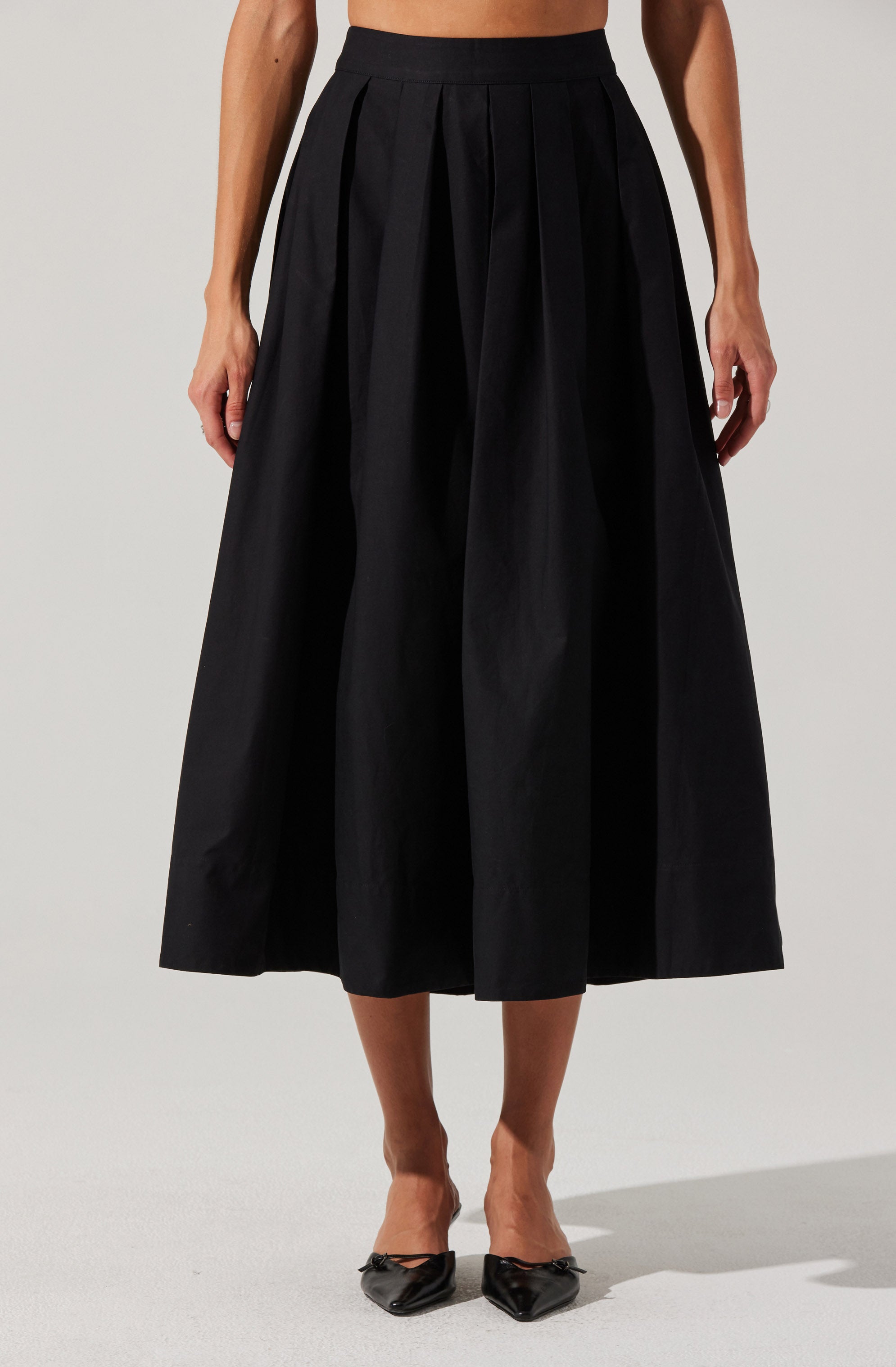 A person is wearing a high-waisted, wide-leg black skirt with pleats, paired with black pointed-toe shoes featuring bows on the front.