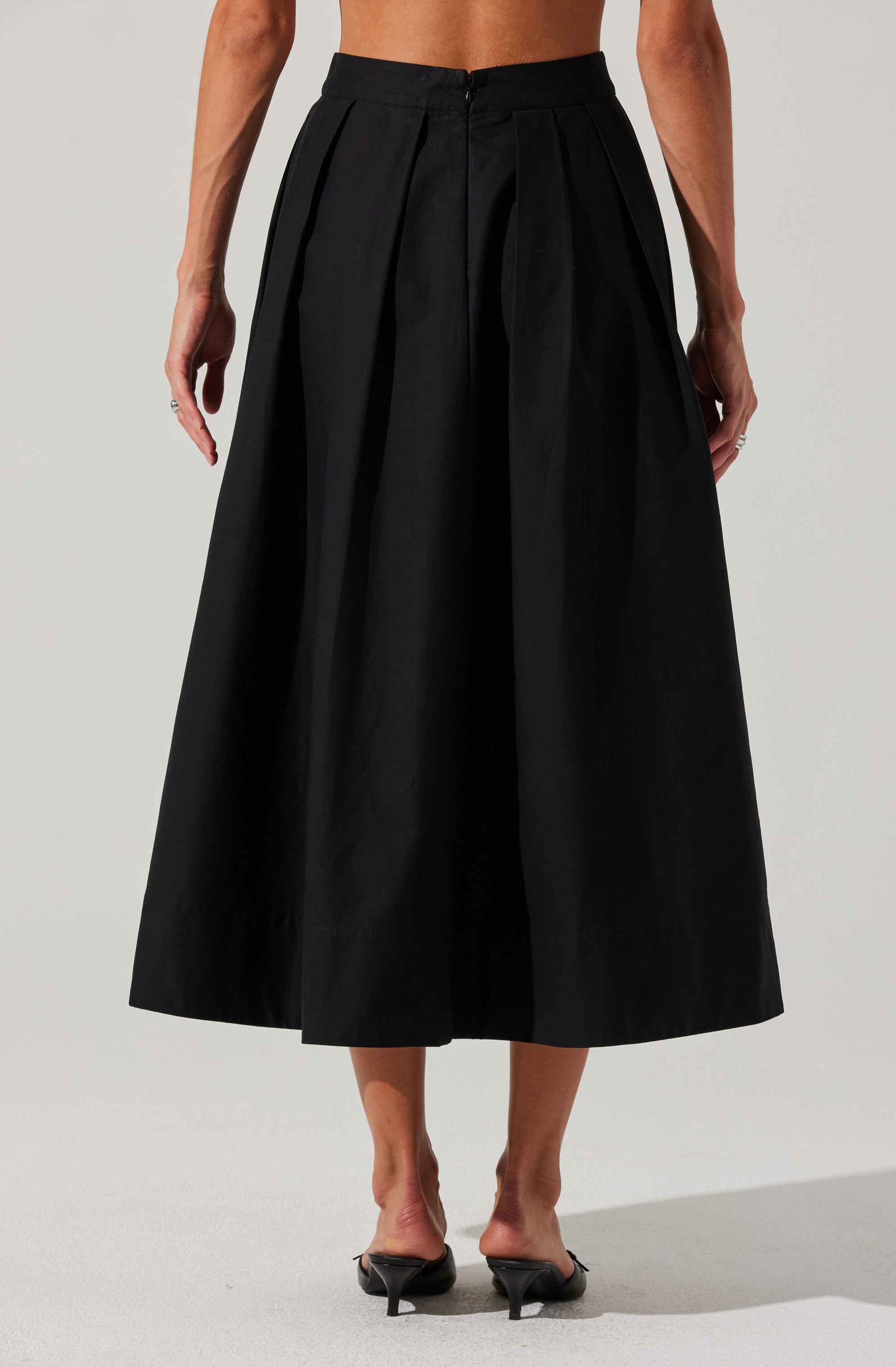 A woman is wearing a high-waisted black midi-length skirt with pleats. She is topless, with her arms relaxed by her sides, and is standing on a plain background. She is wearing black high-heeled shoes.