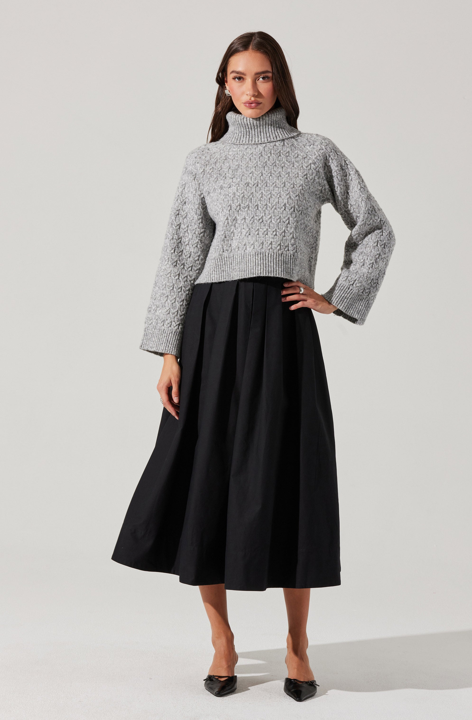 A woman is wearing a light gray, chunky knit turtleneck sweater with a textured pattern. She pairs it with a high-waisted, black, pleated midi skirt and black pointed-toe heels with bow details.