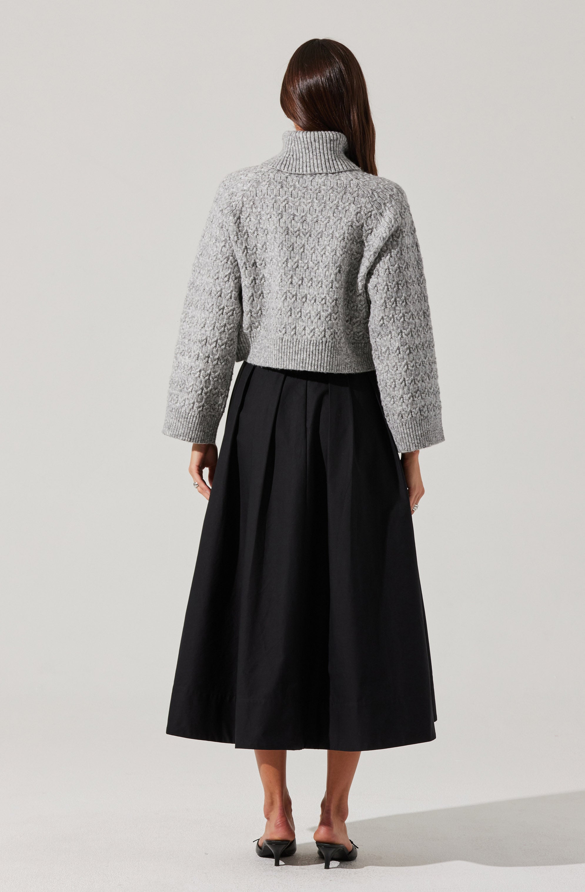 A person is wearing a light gray, chunky knit turtleneck sweater with a textured pattern, paired with a high-waisted, wide-leg black skirt that reaches mid-calf. They are also wearing black high-heeled shoes with ankle straps.