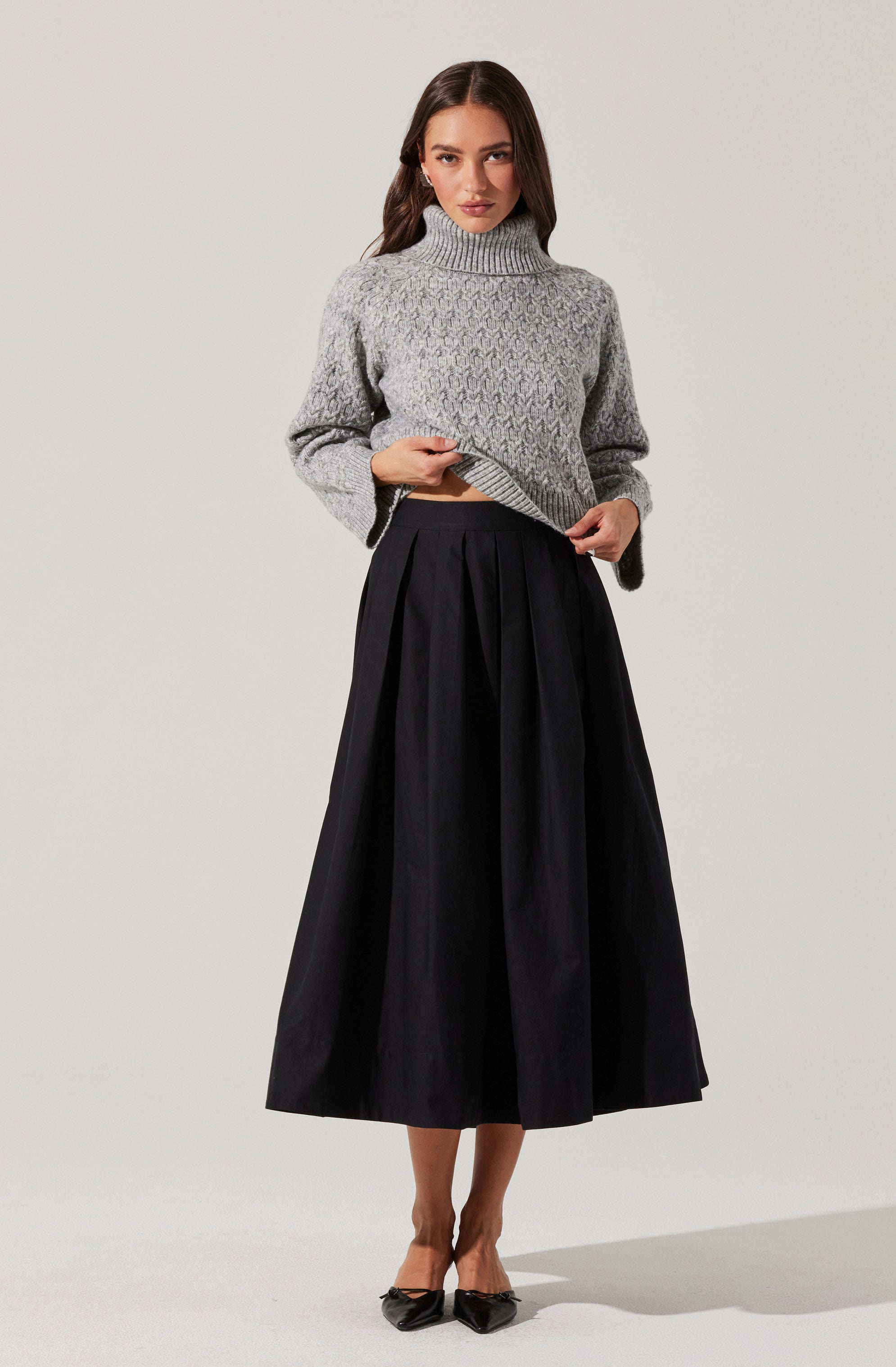 A woman is wearing a gray, cable-knit turtleneck sweater with a cozy, textured pattern. She pairs it with a long, black pleated skirt that reaches mid-calf, creating an elegant and sophisticated look. She completes her outfit with black pointed-toe heels.