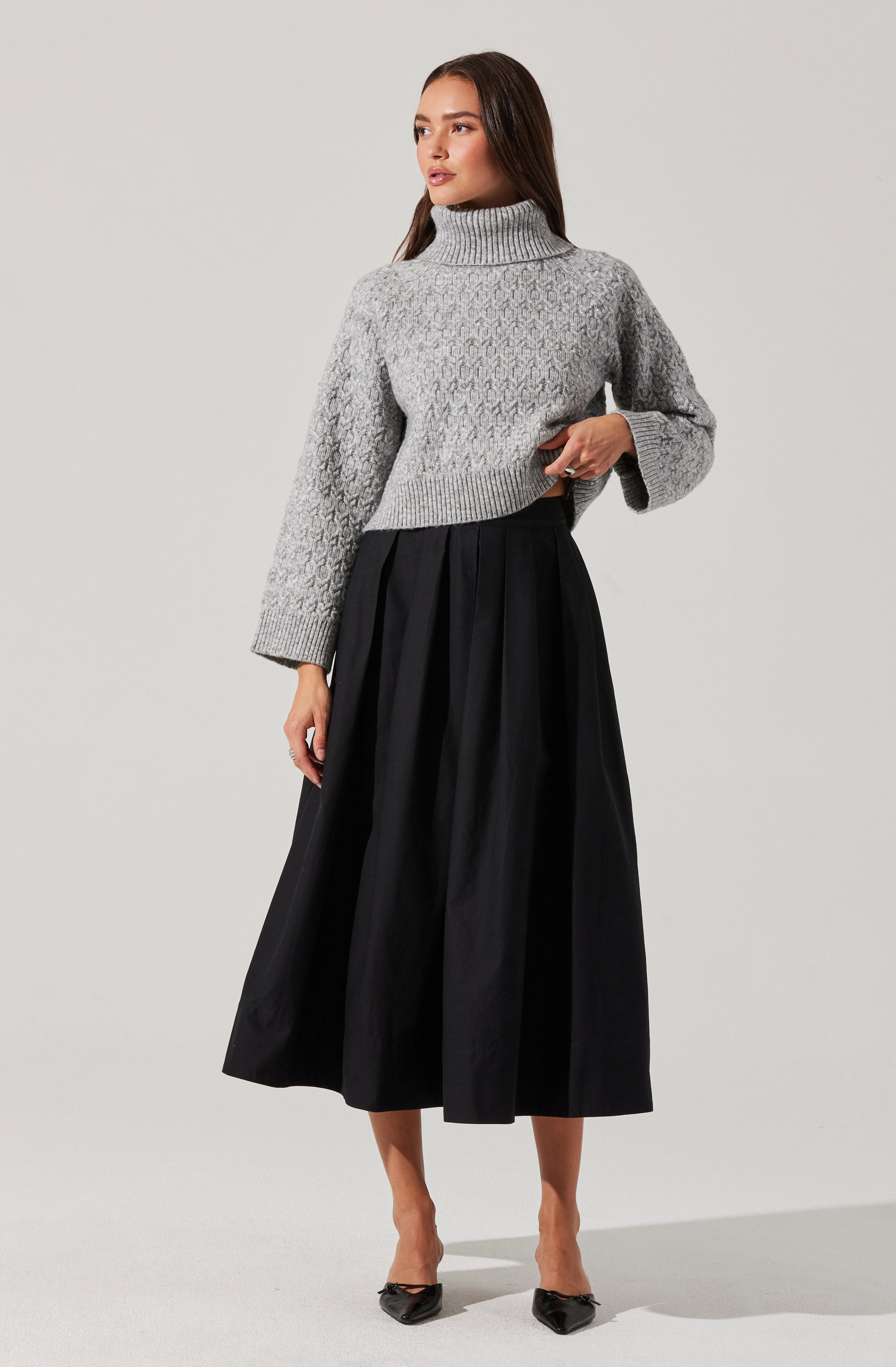 A woman is wearing a light gray, chunky knit turtleneck sweater with textured patterns and long, slightly oversized sleeves. She is pairing it with a high-waisted, black, pleated midi skirt that reaches mid-calf and black pointed-toe high heels.