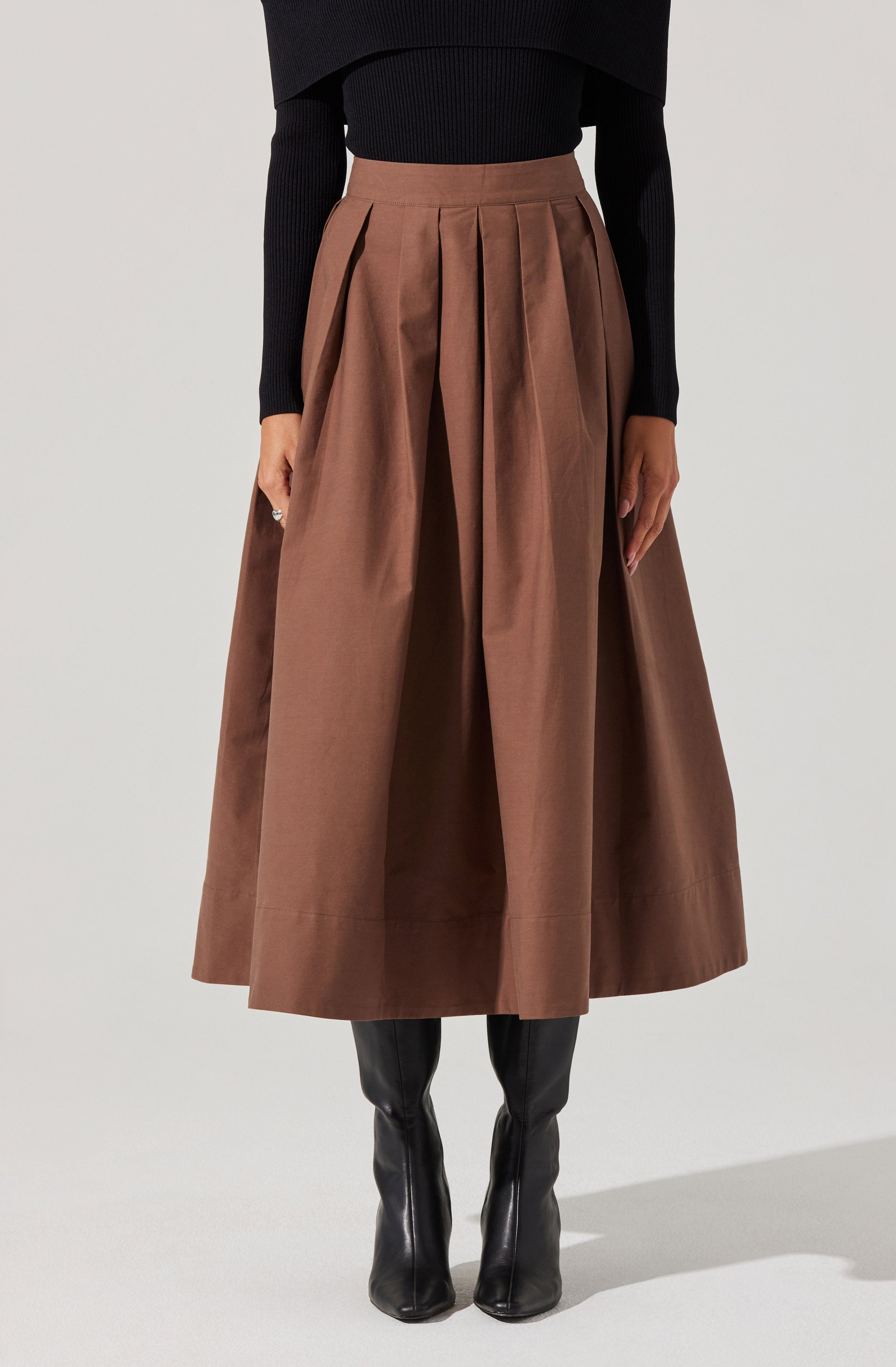 A woman is wearing a high-waisted, wide-leg brown skirt with pleats that extend from the waistband down that give it volume and structure. She pairs the skirt with a long-sleeved, fitted black top that appears to be made of a ribbed fabric. She is also wearing tall black leather boots that reach below the knees.