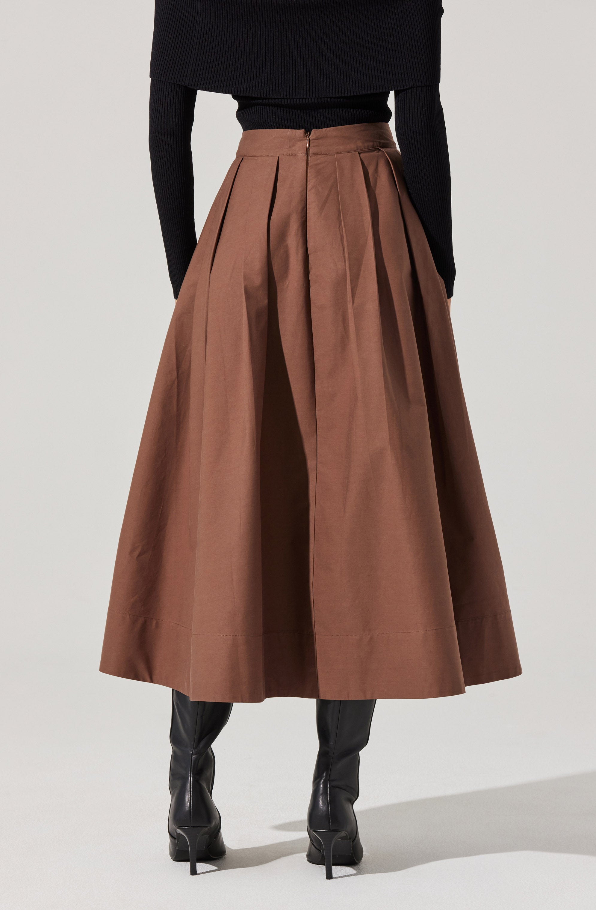 A person is wearing a high-waisted, ankle-length brown skirt with wide pleats. The skirt has a front zipper closure and appears to be made of a sturdy, woven fabric. The person is also wearing a fitted black top with long sleeves.