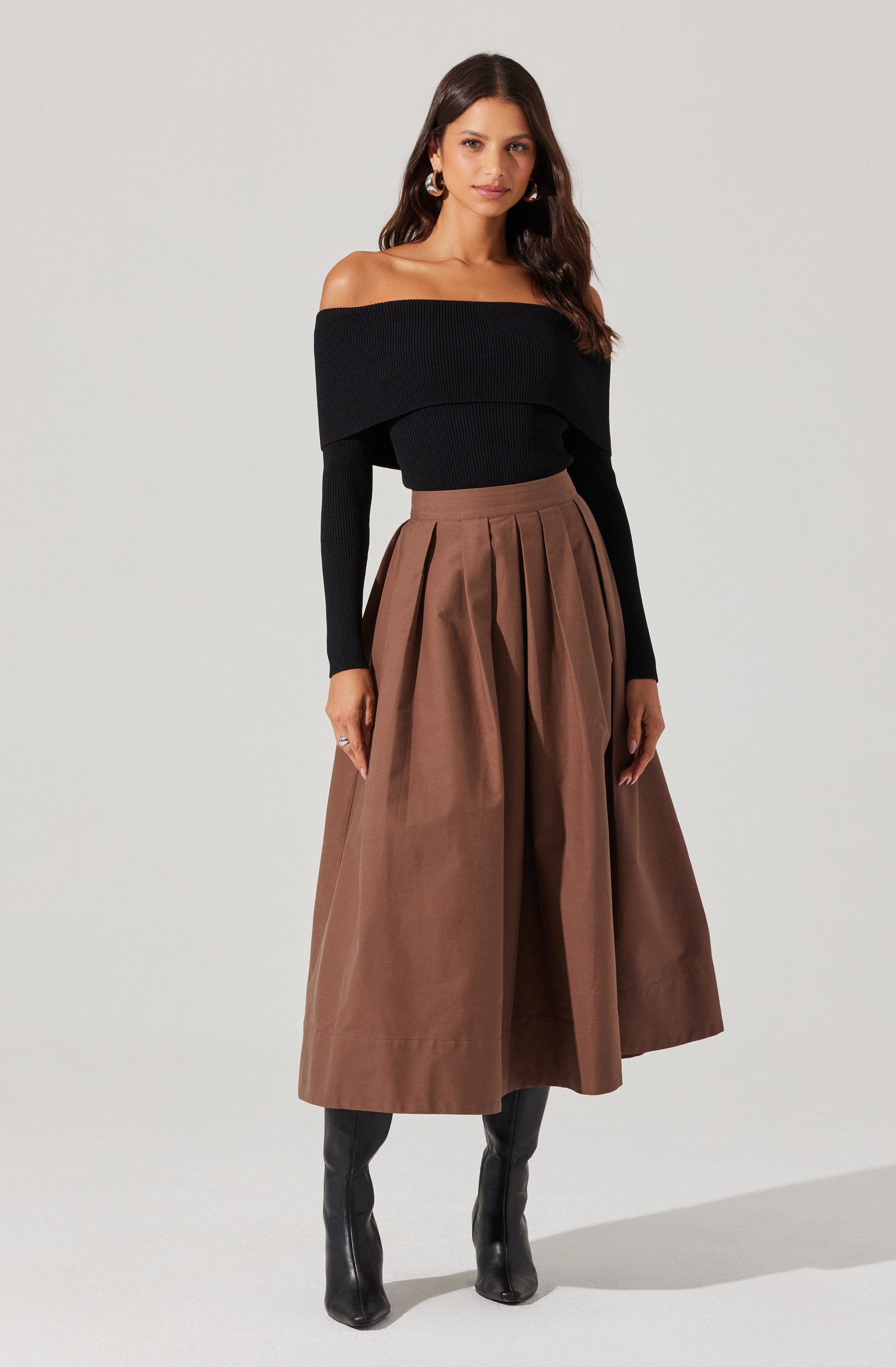 A woman is wearing a black off-the-shoulder sweater with long sleeves and a wide, folded neckline. She pairs it with a high-waisted, A-line midi skirt in a brown, leather-like material with pleats running down the front. She completes her outfit with black knee-high boots.