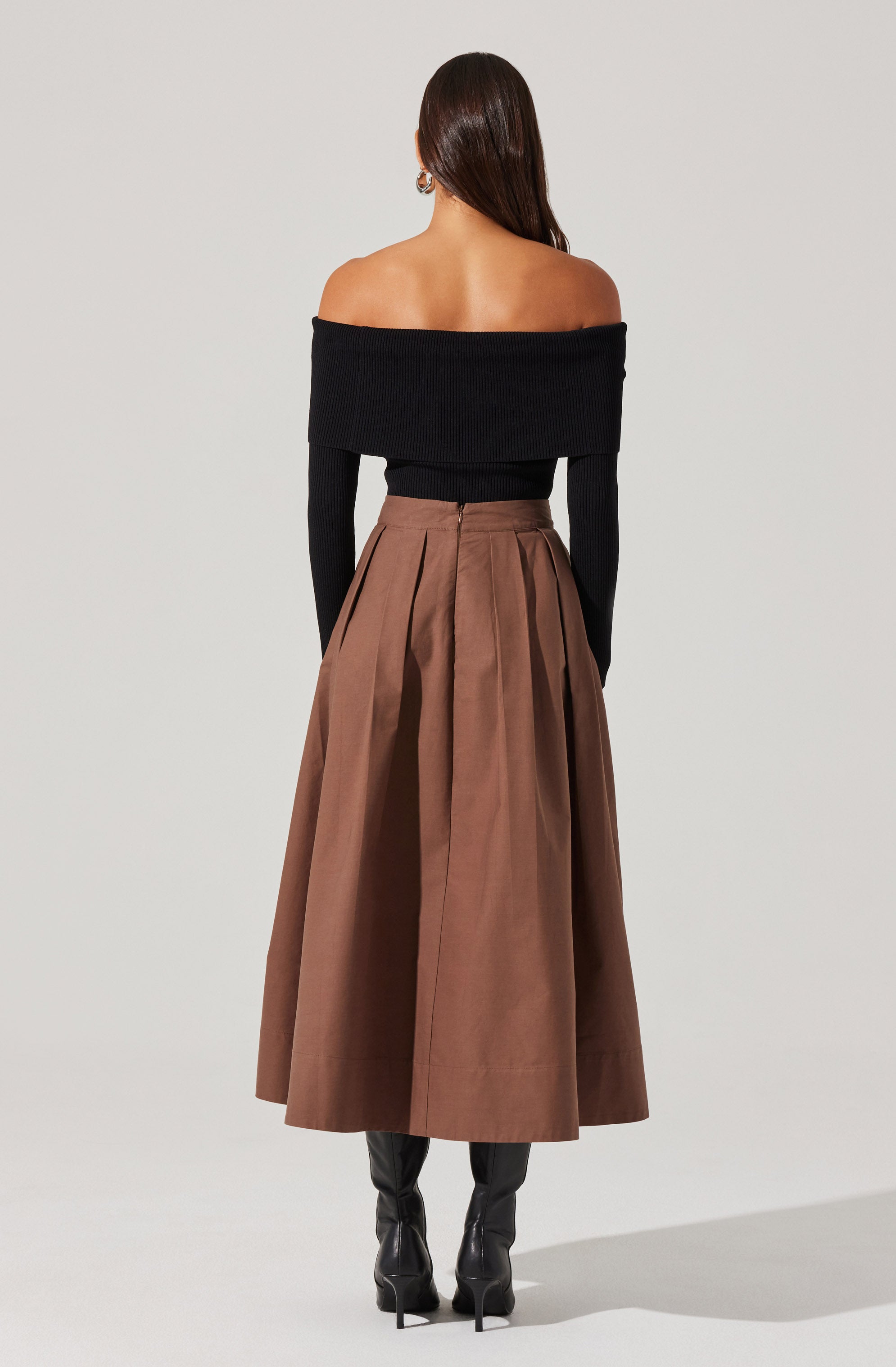 A woman is wearing an off-the-shoulder, long-sleeved black sweater that is fitted at the waist. She pairs it with a high-waisted, full-length brown skirt with pleats and a visible zipper at the back. She completes her outfit with black knee-high boots.